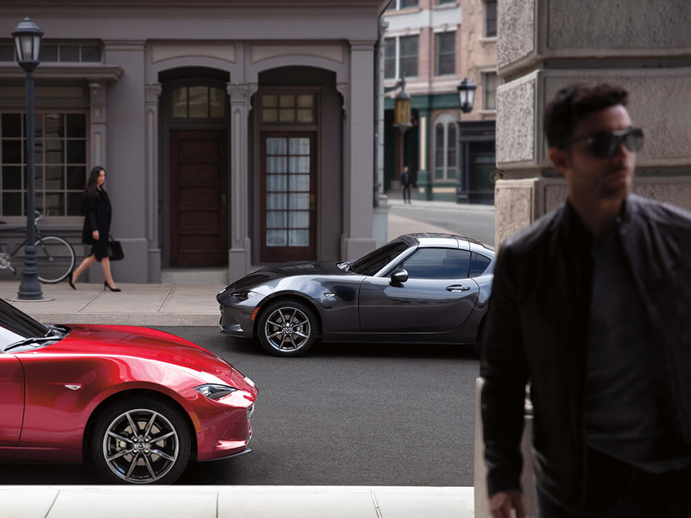 Two gleaming Mazda MX-5 RFs parked along urban street, a woman looks in the background as a man walks away in the foreground.