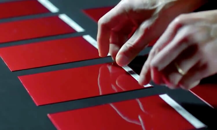 Gros plan de mains qui choisissent un échantillon de peinture rouge sur une table noire sous une lumière intense, parmi d’autres échantillons de couleur similaire.