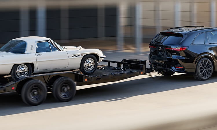 Jet Black Mica CX-70 towing a vehicle trailer hauling a white, classic Mazda leisure vehicle.