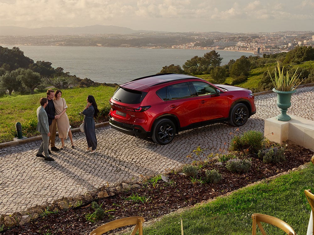 Profil arrière de trois quarts d'un Mazda CX-5 rouge vibrant cristal, avec une femme debout derrière le véhicule, et une ville lointaine visible au-delà d'une étendue d'eau en arrière-plan.