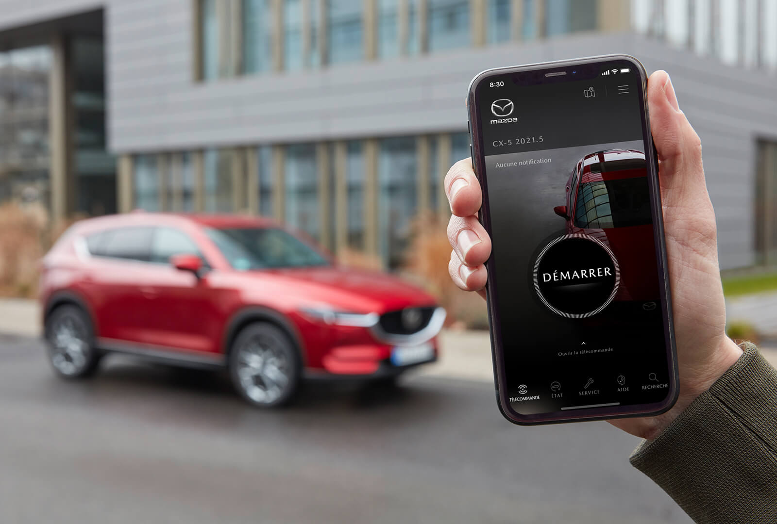 Un homme tient un téléphone affichant l’appli MyMazda tandis qu’un VUS Mazda rouge garé est visible en arrière-plan