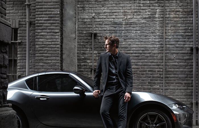 Young professional male wearing dark blazer leans against front passenger fender of Machine Grey Metallic Mazda MX-5 RF parked in industrial alleyway.