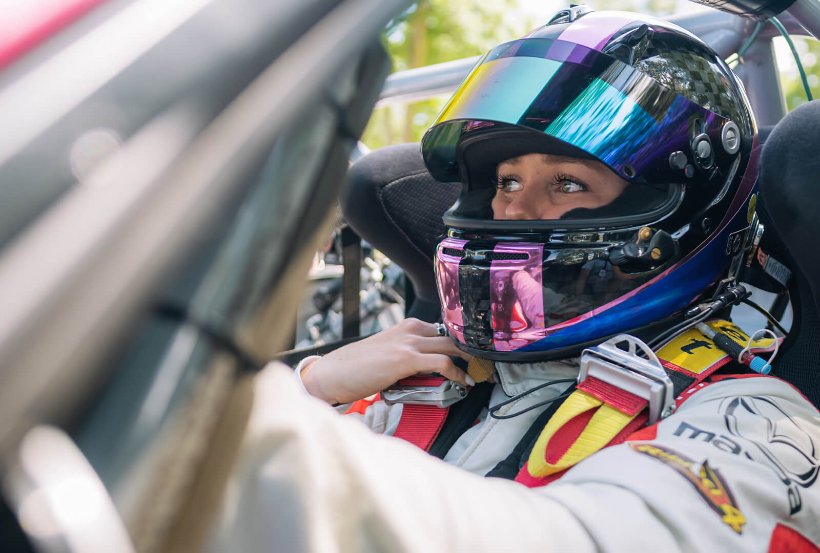 A driver sits behind the wheel getting a safety harness adjusted. She is wearing a driving helmet and full racing gear.