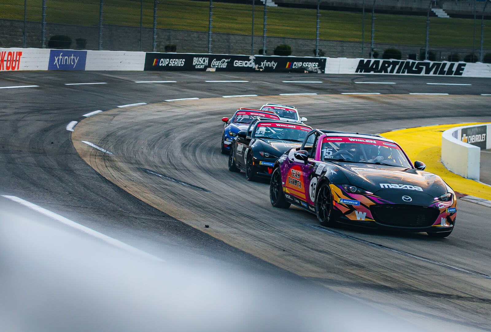 Four racing MX-5s round the corner on a track while driving in single file.