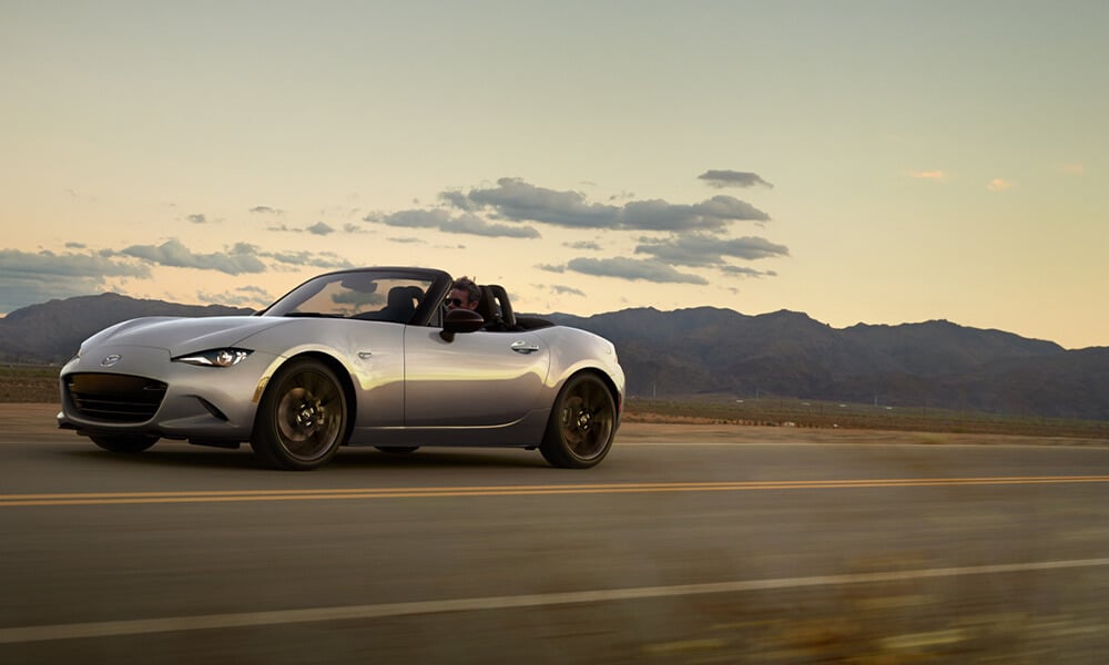 A silver MX-5 with the top down drives down an open road with mountains in the background.