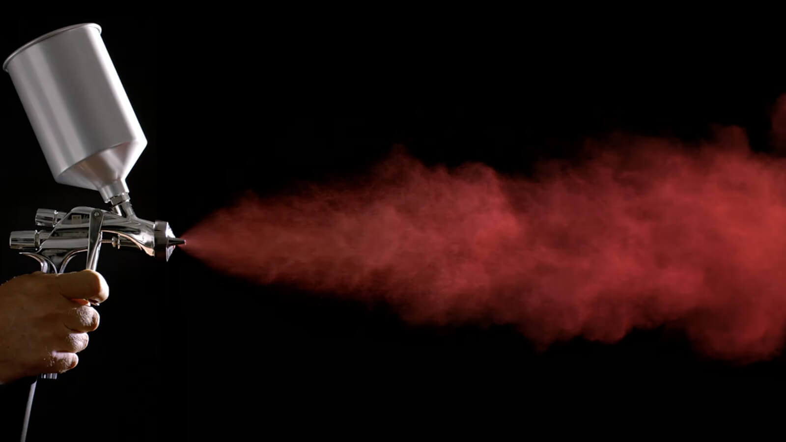 Hand holding spray gun shoots cloud of fine red mist against a dark background