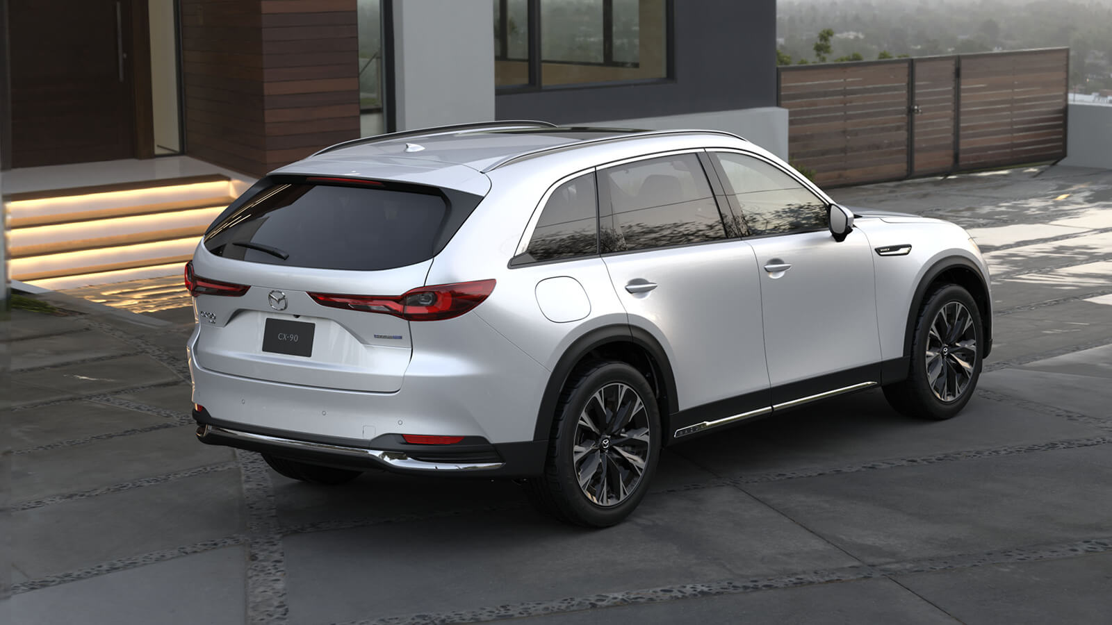 Rear view of CX-90 PHEV, parked in the driveway of building that overlooks a lush, verdant valley in daylight. 