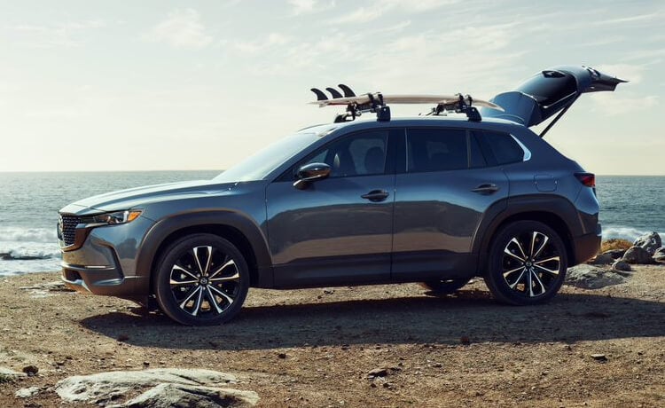 CX-50 with roof rack holding a surfboard, backliftgate is ajar atop a small hill near a beach. 