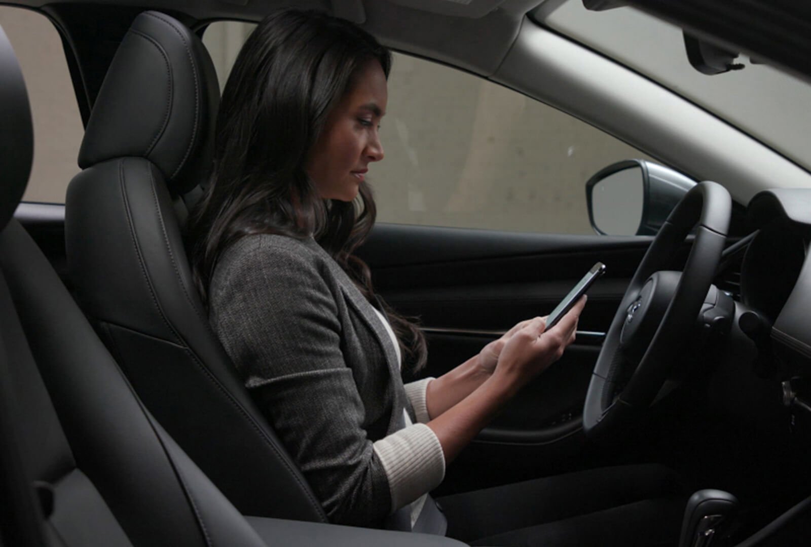 Interior view of woman in driver’s seat of parked Mazda SUV looking at smartphone she’s holding in her hands.
