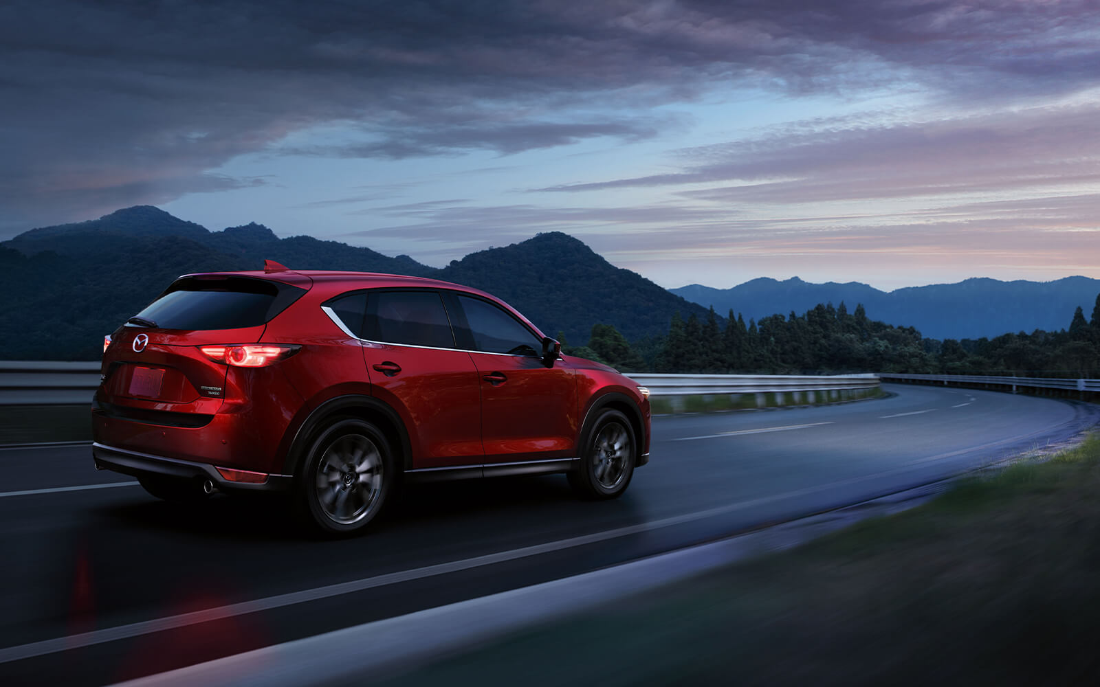 Soul Red Mazda SUV with lights on rounds highway curve at twilight with dark mountains ahead in the distance. 