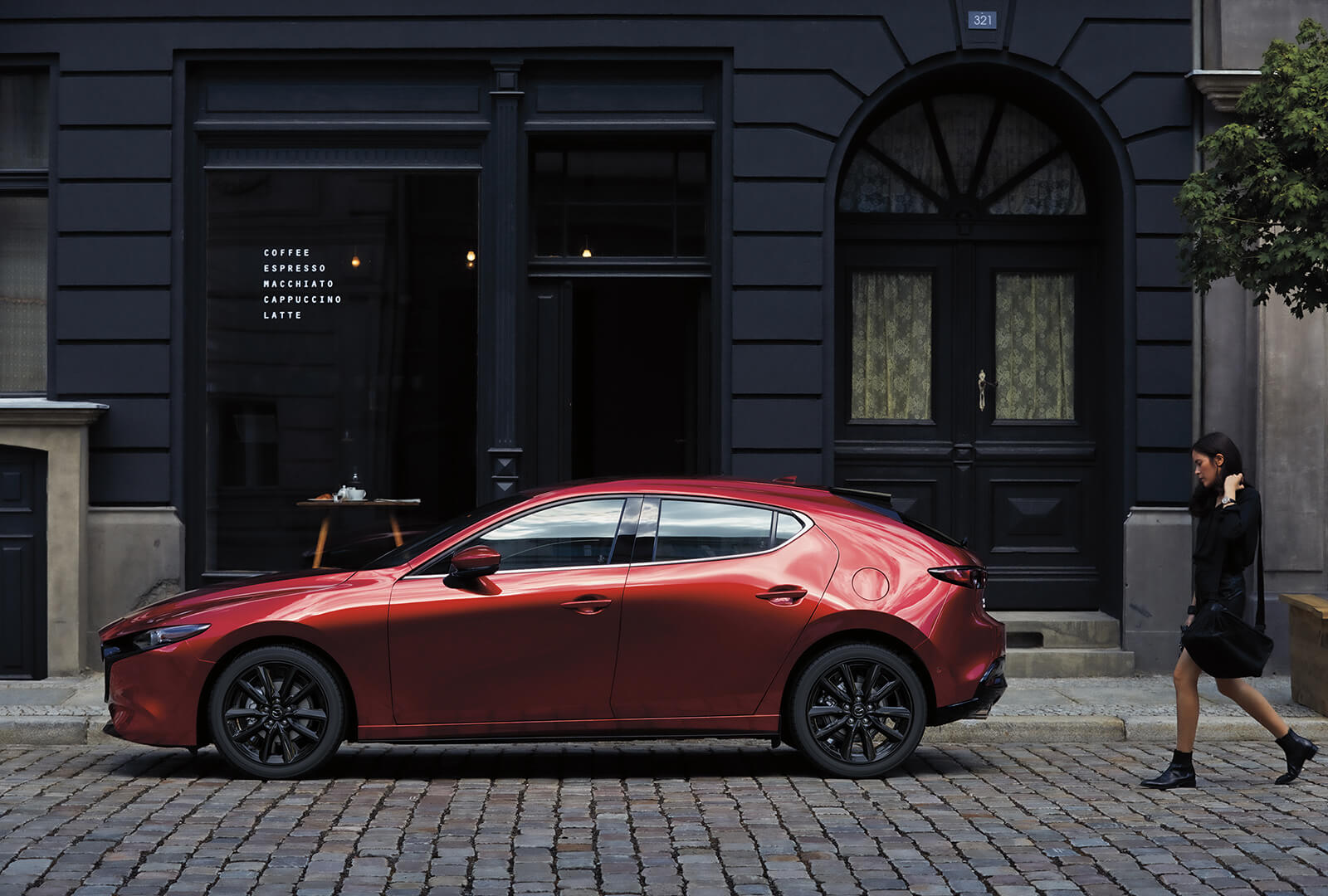 Red Mazda3 side view on cobblestone street with fashionable young woman approaching from right side