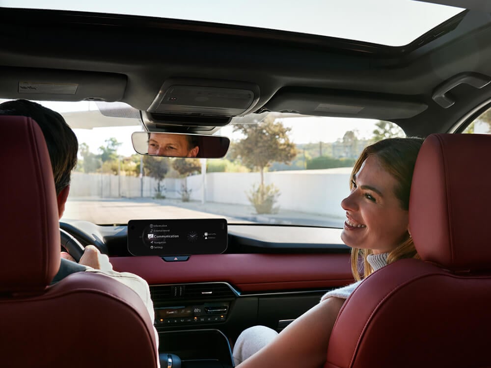 From back seat looking forward in a Mazda CX-70 MHEV with stylish Garnet Red Nappa Leather interior. A woman in the passenger seat is turning to look back. A man is driving, his eyes are visible in rear view mirror.