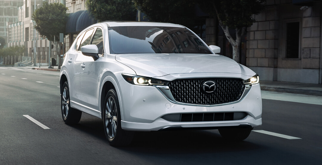 ¾ front image of Rhodium White Metallic Mazda CX-5 driving on city street.