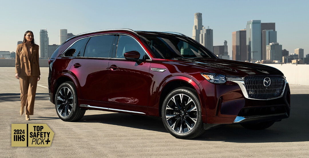 ¾ front shot of parked Artisan Red Mazda CX-90 MHEV parked, cityscape wide in background. A woman walks beside the vehicle.