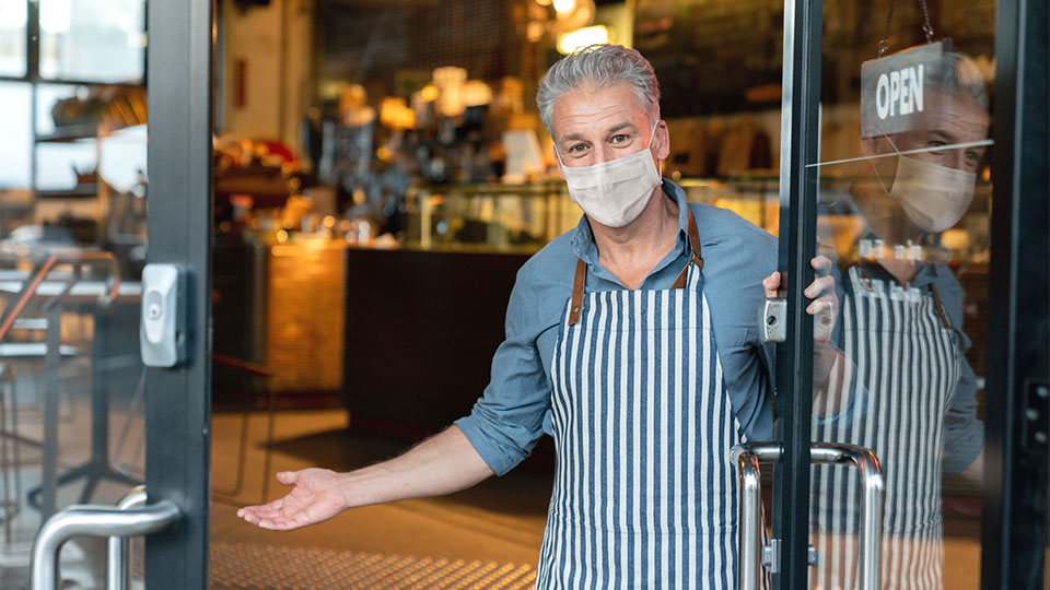 A man in a facemask and striped apron welcomes you into his bakery.  