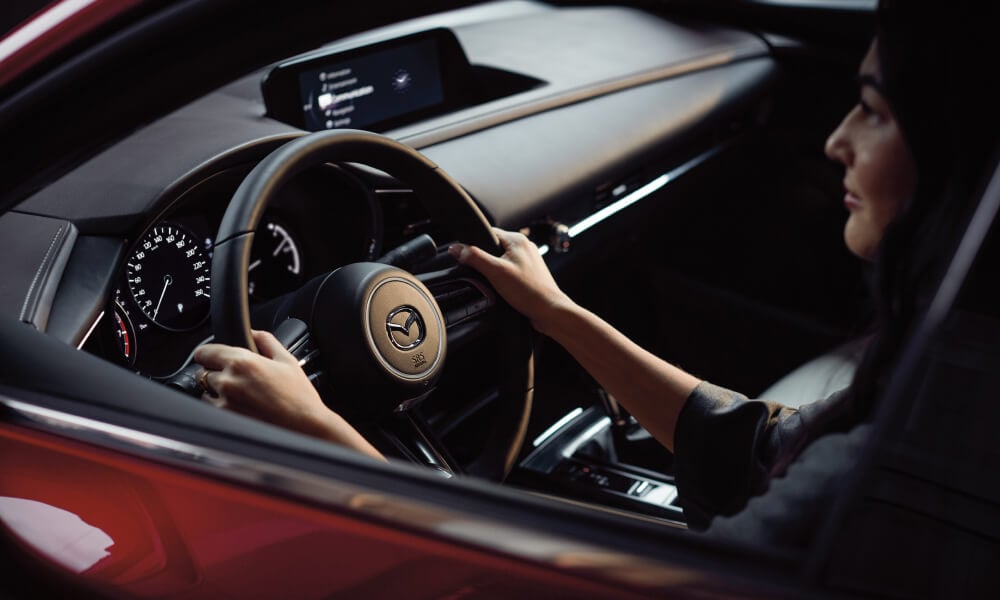 Interior over-the-shoulder shot of woman with hands at 10 and 2 on the wheel with the Mazda logo catching the light.