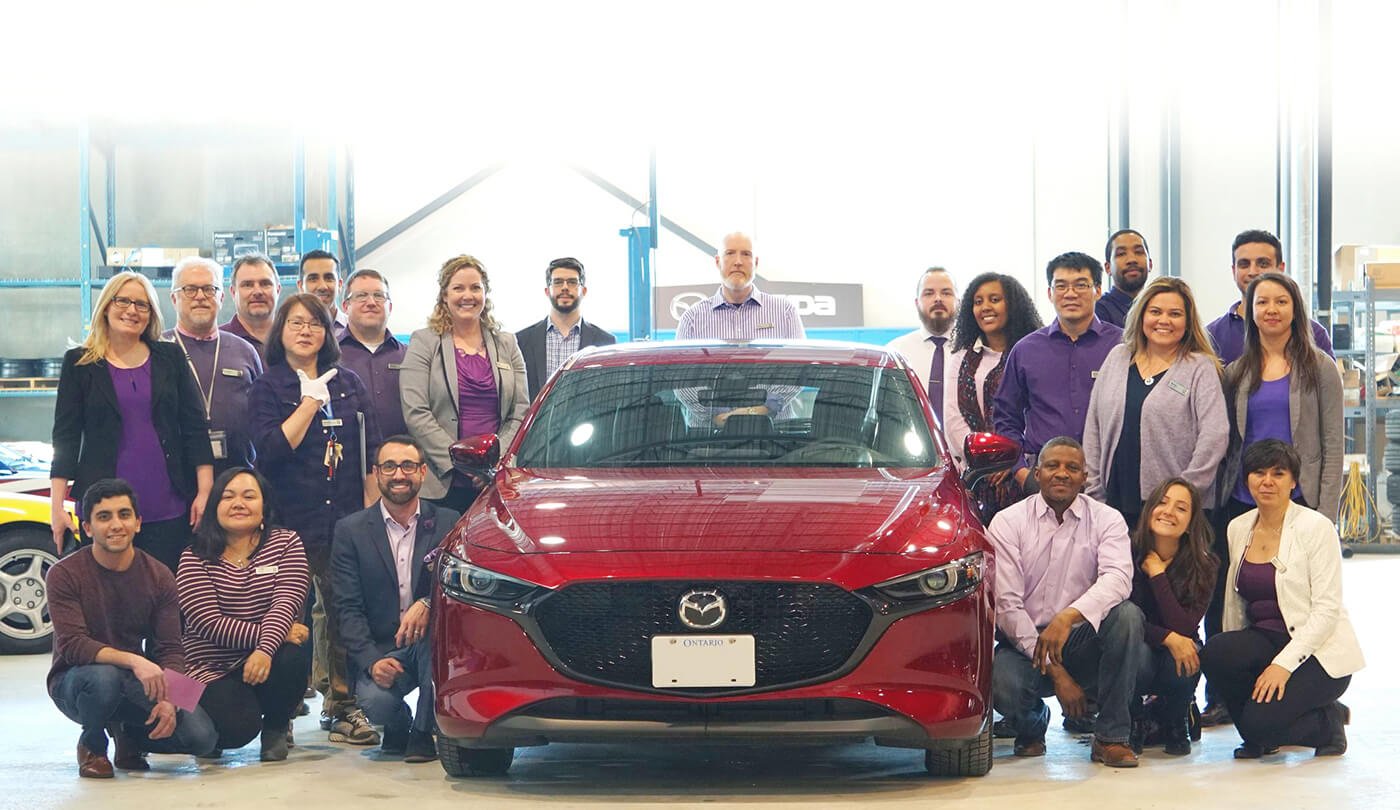 Group photo of Mazda retailer employees standing or kneeling on both sides of a red Mazda3