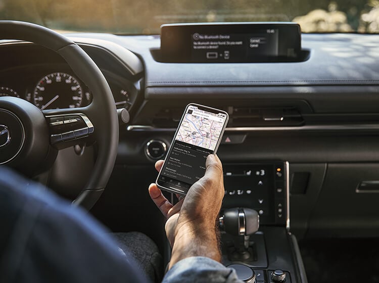 Vue par-dessus l’épaule d’un conducteur qui tient un téléphone affichant une carte, avec l’écran Mazda Connect en arrière-plan.