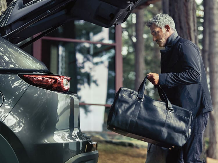 Un homme barbu aux cheveux poivre et sel portant un blouson en cuir et un sac de voyage s’approche d’un VUS Mazda au hayon ouvert
