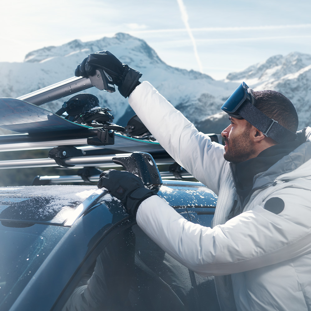  Un homme habillé pour le ski, qui attache un ski sur le toit de sa Mazda CX-50, au sommet d’une montagne enneigée