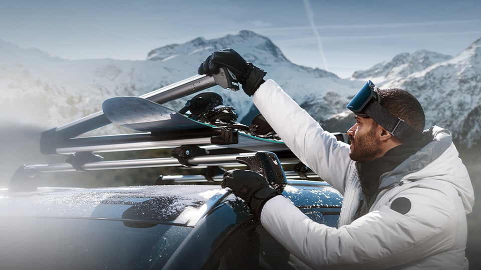 Un homme habillé pour skier tenant un ski près de sa Mazda CX-50 au sommet d’une montagne enneigée.
