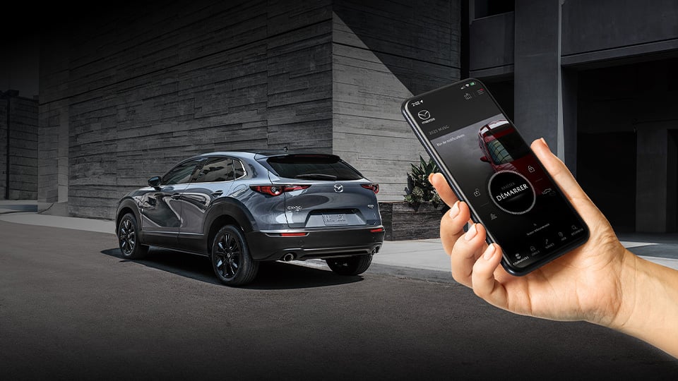 Une main tenant un téléphone sur lequel l'appli MyMazda est ouverte; un CX-30 est présent en arrière-plan