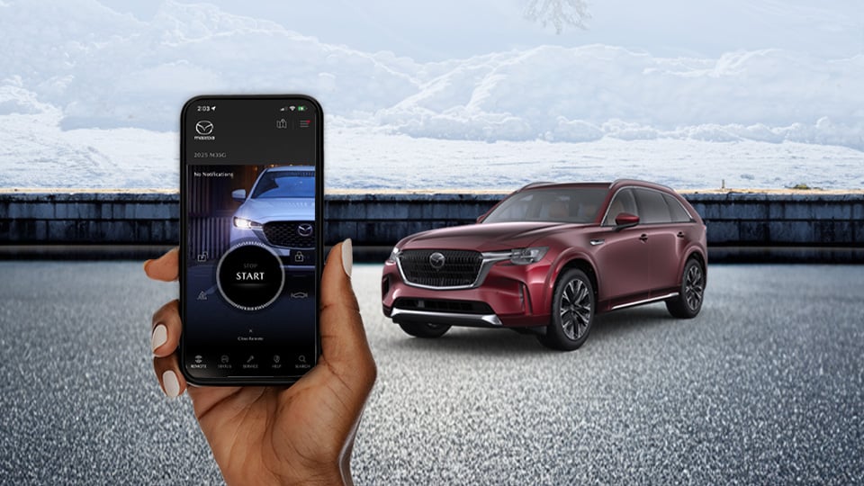 A hand holding a cellphone with the MyMazda app open, with a CX-90 shown in the background
