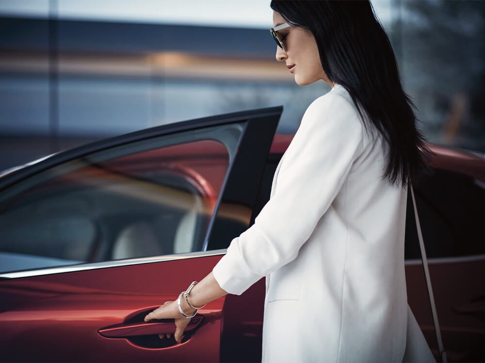 Une femme portant un veston blanc et des lunettes de soleil ouvre la porte d’une Mazda rouge, côté conducteur.