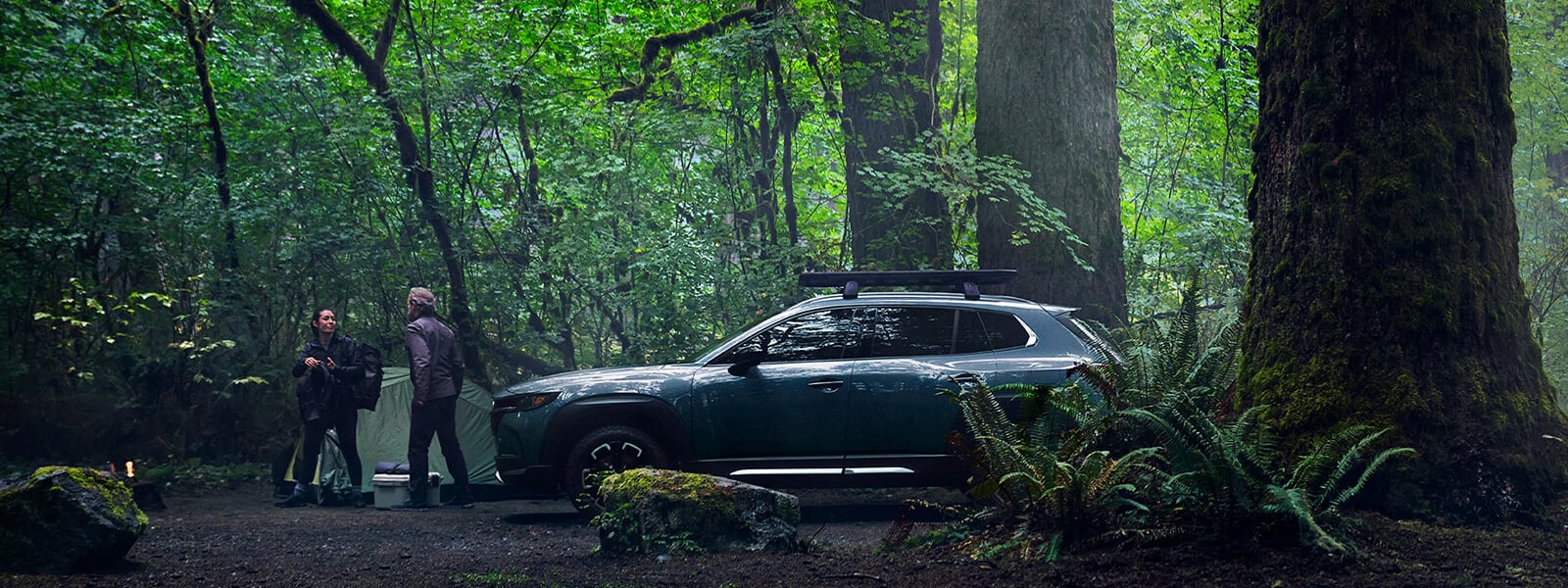 Couple talk between tent and parked Mazda CX-50 amid lush forest foliage and tree trunks.