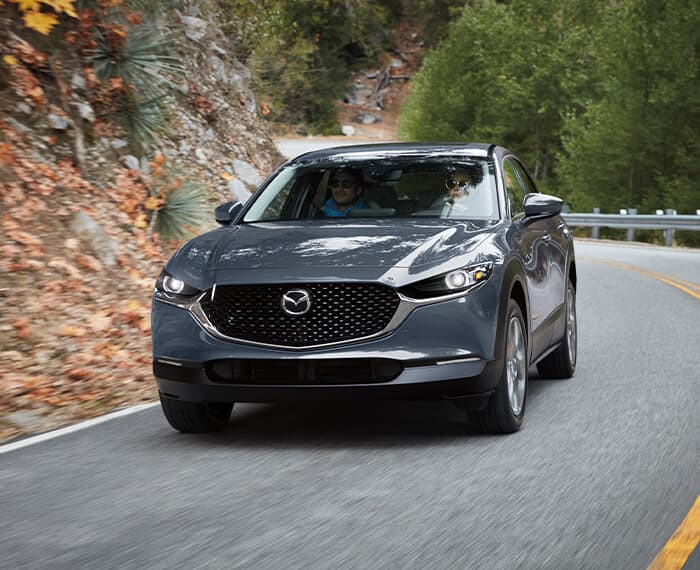 Polymetal Grey Metallic Mazda CX-30 comes out of curve on forested fall highway.