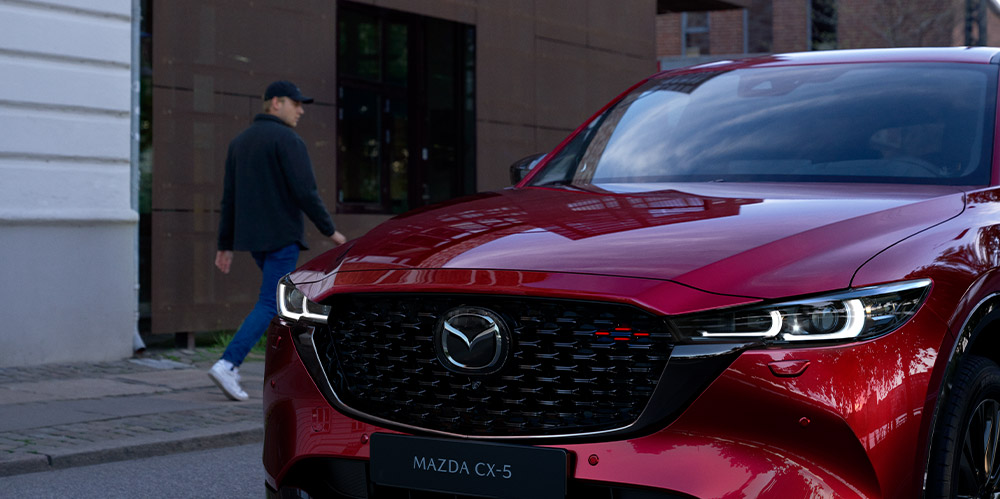 “Soul Red Crystal Metallic Mazda CX-5 with Sport Design trim approaches on urban street.”