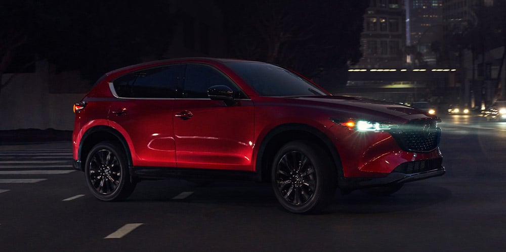 Soul Red Crystal Metallic Mazda CX-5 Sport Design headlights beam as it crosses intersection at night. 