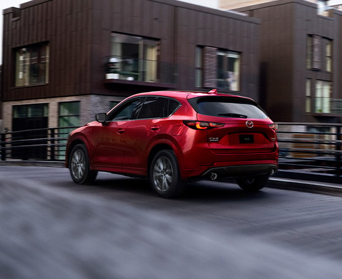 Soul Red Crystal Metallic Mazda CX-5 drives away over bridge past brown-clad buildings.