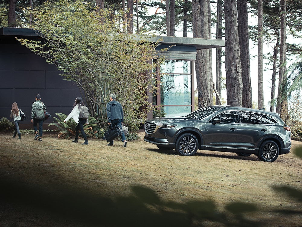 Une famille de quatre personnes décharge leur Mazda CX-9 gris métallisé garée près d'une maison dans les bois.