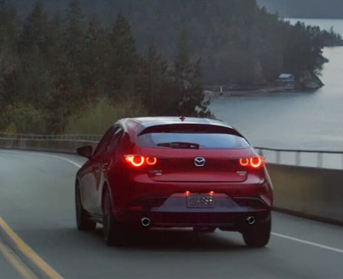 Mazda3 Sport hatchback drives away over bridge with forested coastline ahead in the distance.