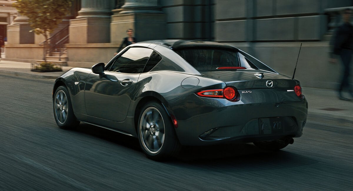 Mazda MX-5 RF with roof up drives away past blurred urban façade and pedestrian in fading light.