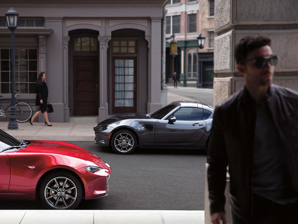 Deux rutilantes Mazda MX-5 RF garées le long d’une rue urbaine; une femme regarde en arrière-plan tandis qu’un homme s’éloigne au premier plan.