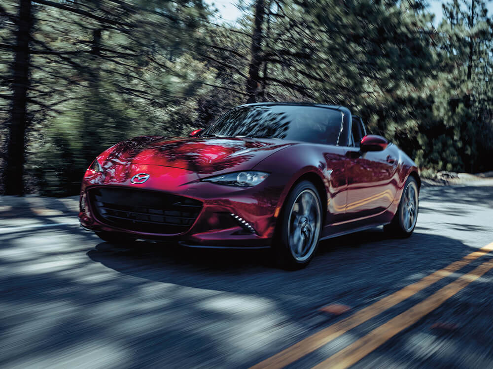 Soul Red Crystal Metallic Mazda MX-5 Soft Top with the top down moves past blurred trees.