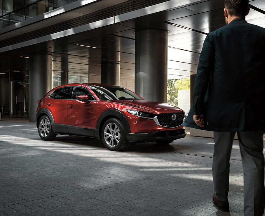 Man approaches Soul Red Crystal Metallic CX-30 parked in the shade of a modern styled building.
