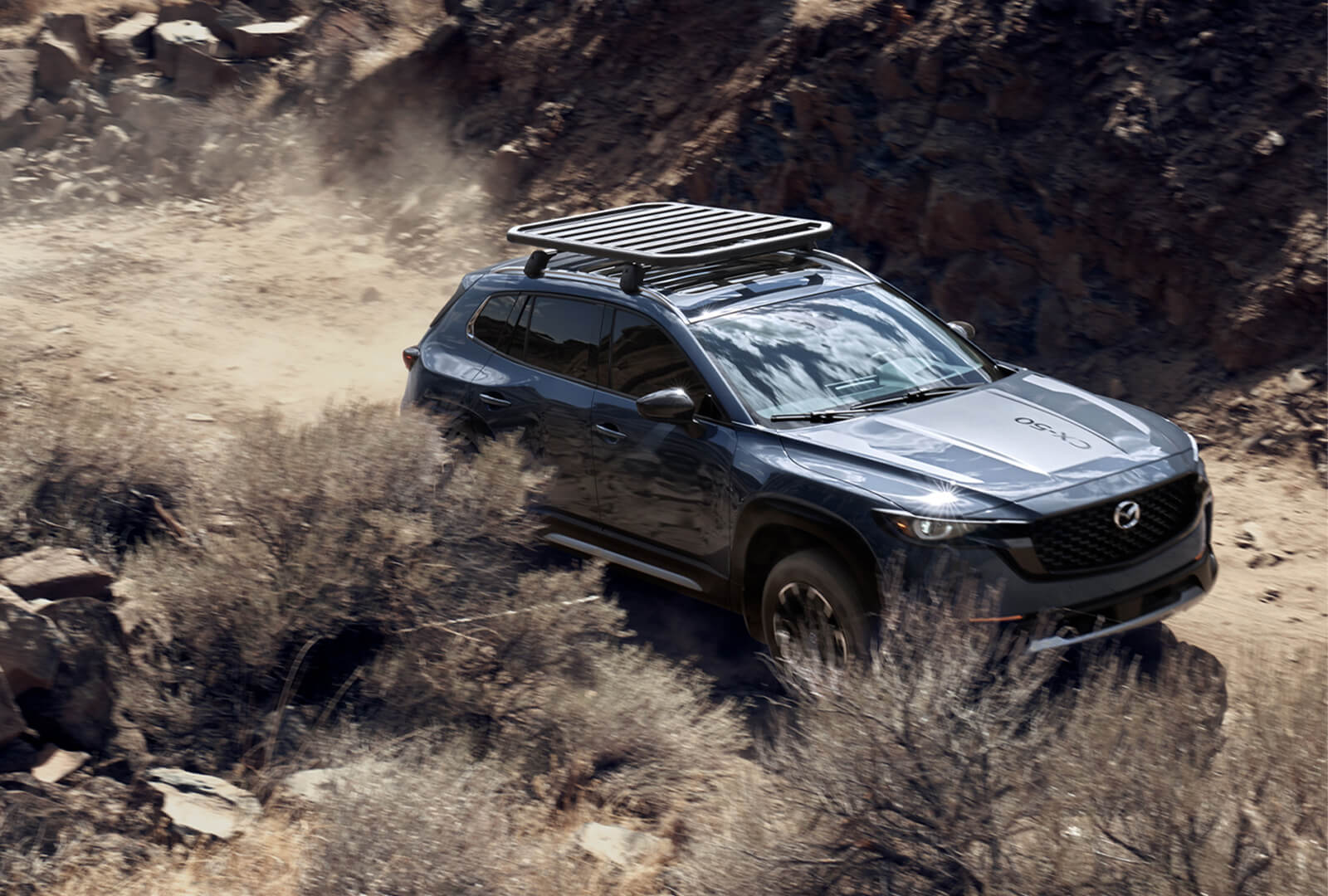 CX-50 with roof rack drives along dusty path.