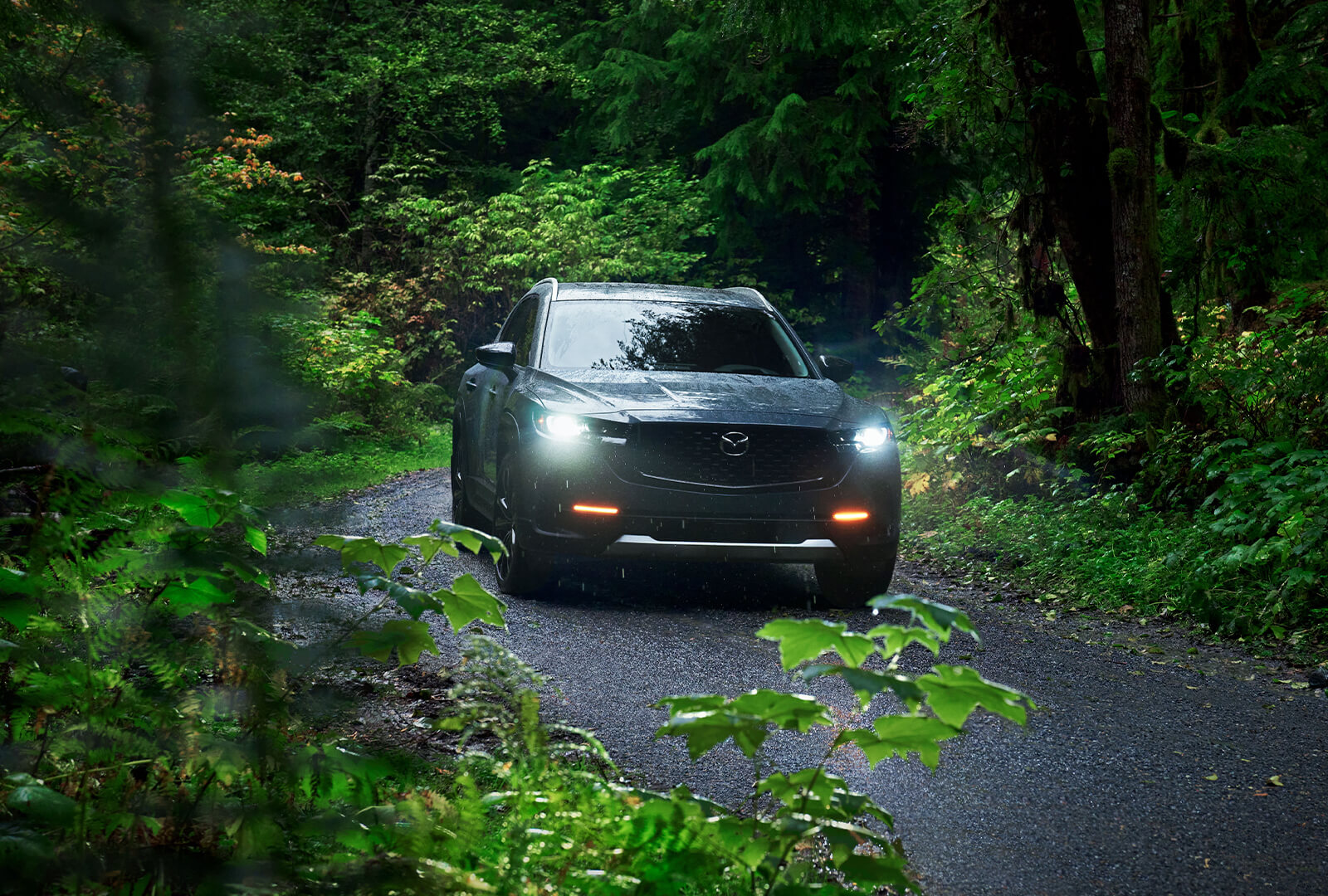 Mazda CX-50 approaches after rounding a curve on a gravel road in dense forest.