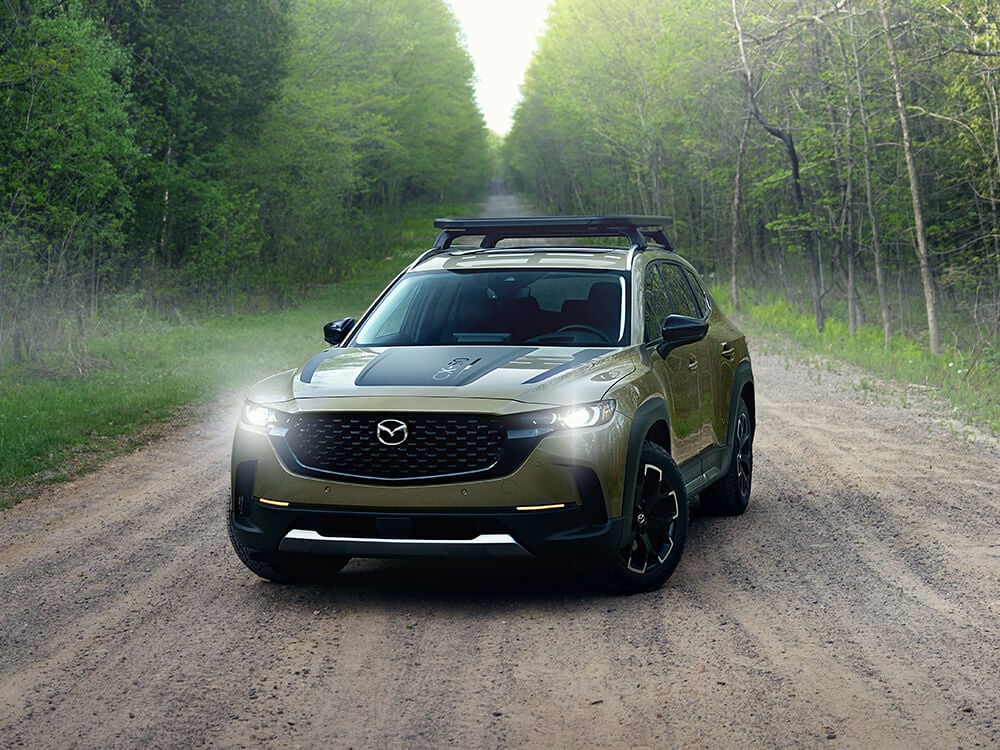 Front view of parked CX-50 with Meridian trim on gravel road. Front lights are on.