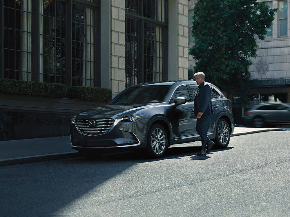 Man in navy blue suit approaches a parked CX-9 on a city street.