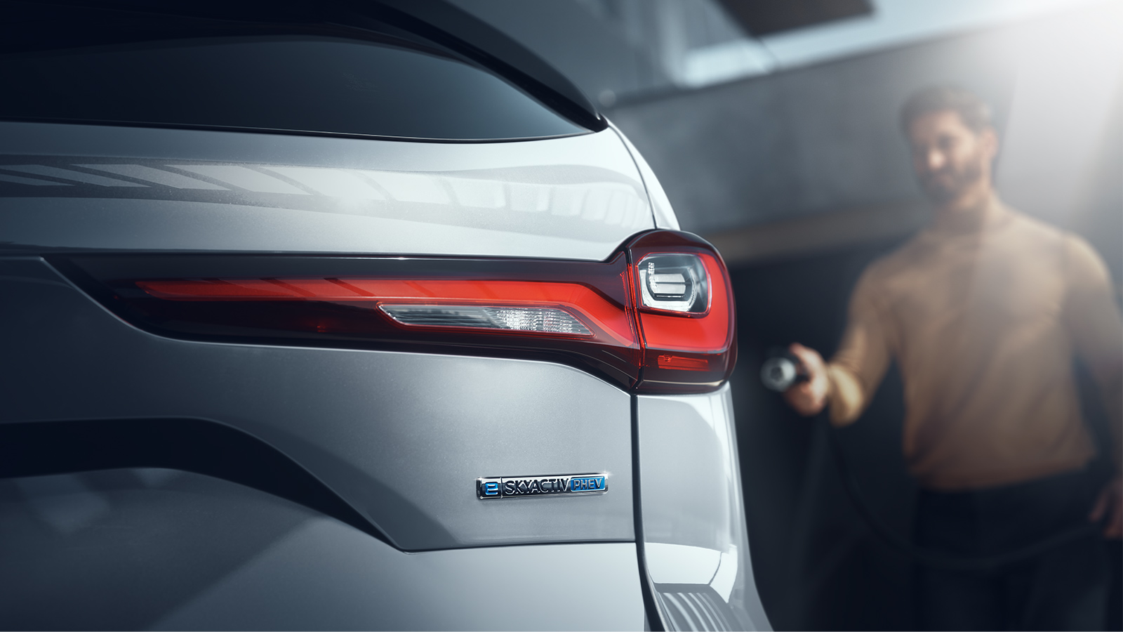 Grey CX-90 exterior closeup with a focus on the PHEV eblem with a blurred person in the back with a plug to emphasize the plug-in feature.