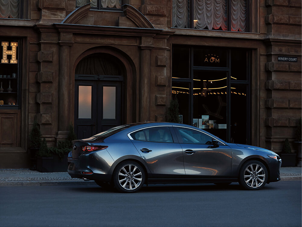 Une Mazda3 gris polymétal métallisé garée devant l’entrée d’un bâtiment.