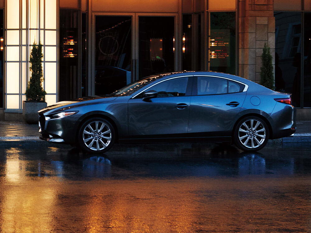 Une Mazda3 gris mécanique métallisé garée devant une salle de spectacle. La chaussée est mouillée et le véhicule reflète les lumières du chapiteau.