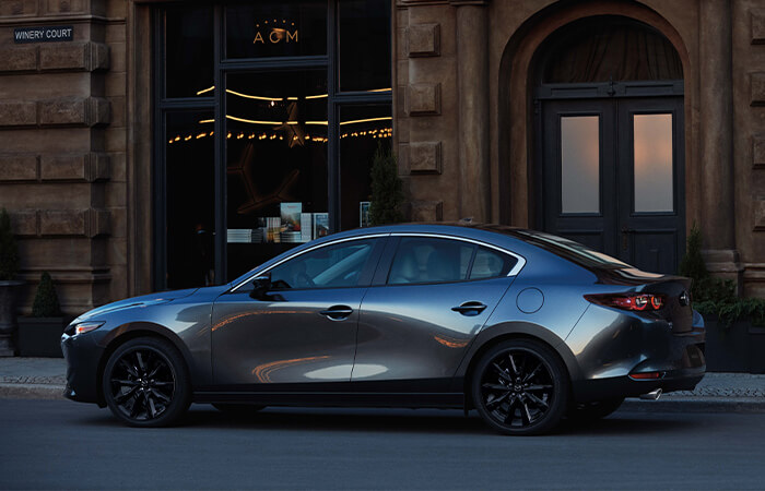 Driver’s side profile of Machine Grey Metallic Mazda3 reflecting light on a city street.