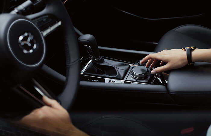 Un conducteur dans l’habitacle de la Mazda3, une main tendue vers le bouton de la console centrale et l’autre sur le volant.
