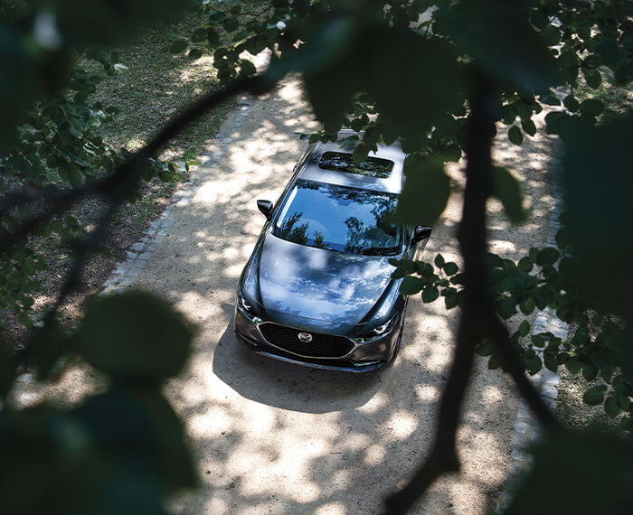 Mazda3 sedan drives along rural road past blurred tree.
