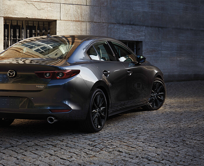 Mazda3 sedan reflects natural light while turning onto a cobblestone road.